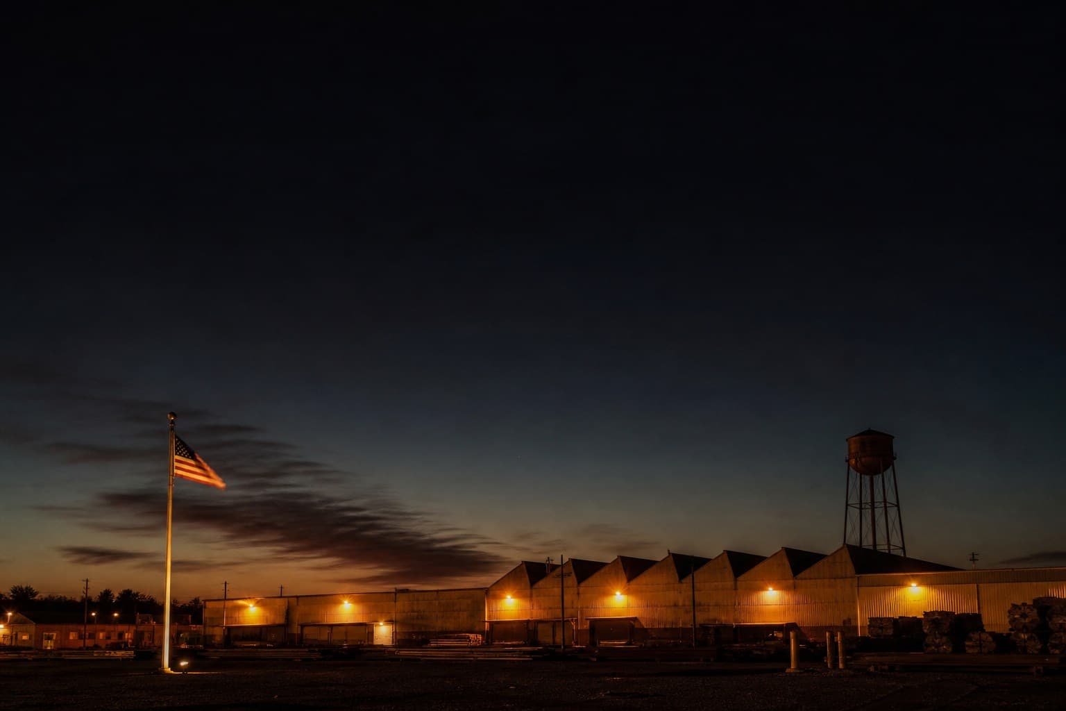 An American factory at dusk: a sawtooth-roof plant with warm lights along its wall, a water tower above the roofline, an American flag on a floodlit pole in the yard